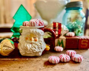 May include: A ceramic mug shaped like a Santa Claus face with a red handle, filled with small round white and red striped peppermint candies. The mug features a sculpted white beard, mustache, and hat brim, with closed eyes and rosy cheeks.