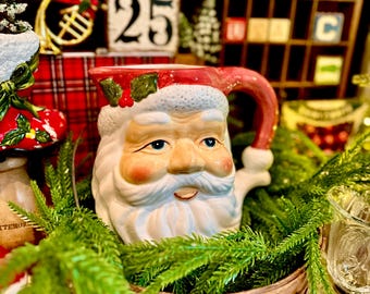 Könnte beinhalten: Ein Keramik-Weihnachtsmannkrug mit roter Mütze, grünen Stechpalmenblättern, roten Beeren, weißem Bart und einem Henkel. Der Krug steht in einem Holzkorb, der mit künstlichen grünen Tannenzweigen gefüllt ist.