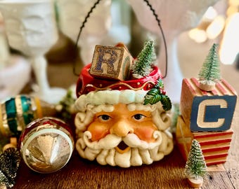 Könnte beinhalten: Ein Keramikbehälter in Form eines Weihnachtsmannkopfes mit schwarzem Drahthenkel und roter Mütze mit Stechpalmenblättern. Im Behälter befinden sich Holzklötze mit dem Buchstaben R und ein kleiner grüner Tannenbaum. Ein Klotz mit dem Buchstaben C steht daneben.