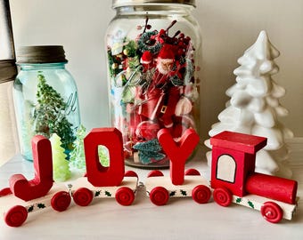 May include: A wooden toy train set with red letters spelling JOY on individual flatbed cars, followed by a red locomotive engine. The train cars feature small red wheels and a white base with green holly leaf accents, displayed with glass jars.