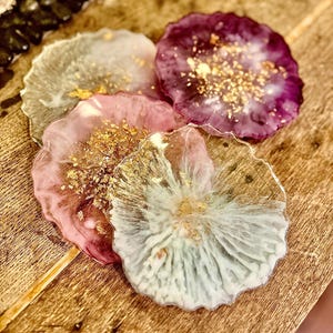 May include: Four resin coasters in shades of pink, purple, and white with gold flecks. The coasters have a unique, organic shape and are arranged on a wooden surface. The coasters are a decorative and functional home decor item.