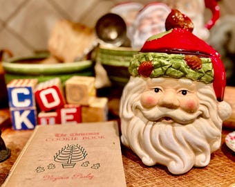May include: A ceramic Santa Claus cookie jar with a red hat and green holly trim, positioned next to a vintage book titled The Christmas Cookie Book by Virginia Pasley and several wooden alphabet blocks spelling out the word COOKIE.