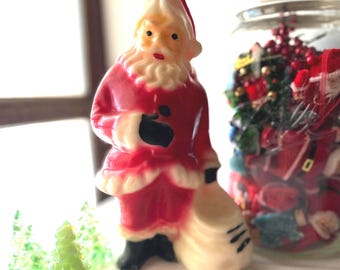 May include: A molded wax Santa Claus figurine wearing a red suit with white trim, black boots, and a black belt, carrying a large white sack. The figure has a white beard, a red hat, and a small metal loop at the top for hanging as an ornament.