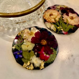 Op de afbeelding: Twee hars coasters met ingebedde gedroogde bloemen in rood, wit en geel, met groene bladeren en gouden vlokken. De coasters hebben een donkere achtergrond. Een heldere glazen kom met een gouden rand staat op de achtergrond.
