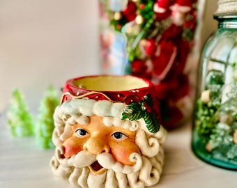 May include: A ceramic Santa Claus head planter featuring a red hat with a white trim, a white beard, and a mustache. The Santa face has rosy cheeks, dark eyes, and a small holly leaf accent on the side of the hat. The interior is cream colored.
