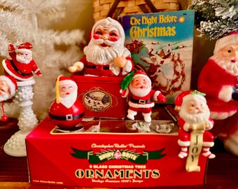 May include: A collection of vintage-style Santa Claus figurines and ornaments arranged around a red box labeled Shiny Brite 9 Glass Christmas Tree Ornaments. A Santa jack-in-the-box, a round Santa ornament, and a book titled The Night Before Christmas.
