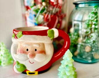 May include: A ceramic mug shaped like a smiling Santa Claus face with a red hat, white beard, and a red handle. The mug features green holly leaves on the brim and a black belt with a yellow buckle at the base, accompanied by small green trees.