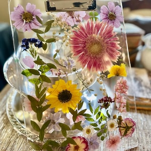 Peut inclure: Un presse-papier en acrylique transparent contenant une variété de fleurs séchées pressées incrustées dans le matériau. La composition comprend une grande marguerite gerbera rose, un tournesol jaune vif et diverses petites fleurs violettes, blanches et roses.