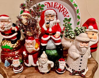 May include: A collection of various Santa Claus figurines in ceramic and glass, featuring red and white suits, some with pipes or sacks. A round white plate with the word MERRY and holly leaves sits in the back, alongside small decorative trees.