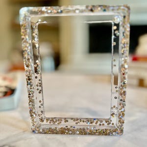 May include: A clear, rectangular picture frame with glitter embedded in the edges. The glitter is a mix of gold, silver, and iridescent colors, creating a sparkling effect. The frame is empty, ready for a photo.