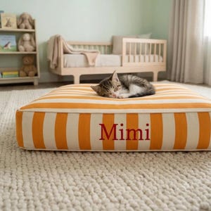 May include: A rectangular pet bed featuring vertical orange and white stripes with the name Mimi embroidered in red on the front side. A small tabby kitten is curled up and sleeping on top of the cushioned surface of the striped pet bed.