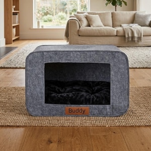 Puede incluir: Una cama rectangular gris para mascotas con una abertura rectangular. El interior tiene un cojín de felpa negro. Una etiqueta de cuero marrón con el nombre "Buddy" está adherida al frente. La cama está colocada sobre una alfombra tejida.