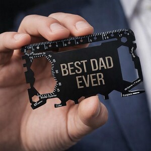 May include: A black multi-tool card with the text "BEST DAD EVER" in silver. The card features a ruler, bottle opener, cutter, and other tools. The card is held in a person's hand.