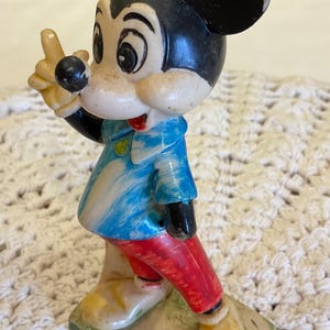 Peut inclure: Une figurine en plastique vintage de Mickey Mouse portant une chemise ray&eacute;e bleu et blanc et un pantalon rouge. La figurine est debout sur un socle avec les mots "Walt Disney Productions" imprim&eacute;s dessus.