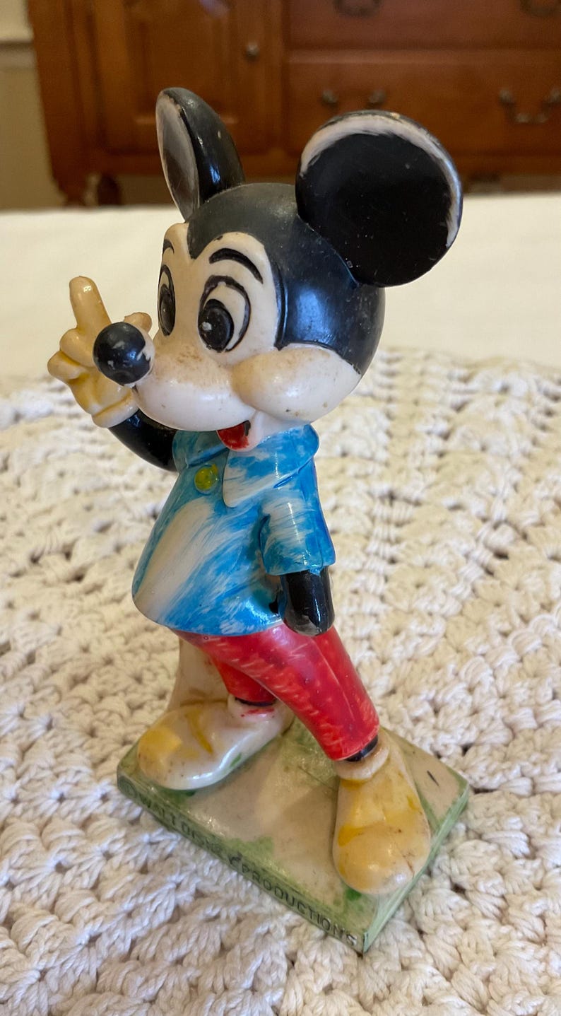 Peut inclure: Une figurine en plastique vintage de Mickey Mouse portant une chemise ray&eacute;e bleu et blanc et un pantalon rouge. La figurine est debout sur un socle avec les mots "Walt Disney Productions" imprim&eacute;s dessus.