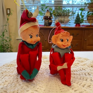 May include: Two vintage Christmas elf figurines with red and green clothing. The elves have plastic heads and are sitting with their legs crossed.