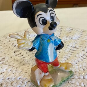 Peut inclure: Une figurine vintage en plastique de Mickey Mouse portant une chemise bleue, un pantalon rouge et des chaussures jaunes. La figurine est debout sur une base verte avec le texte "WALT DISNEY PRODUCTIONS" en bas.