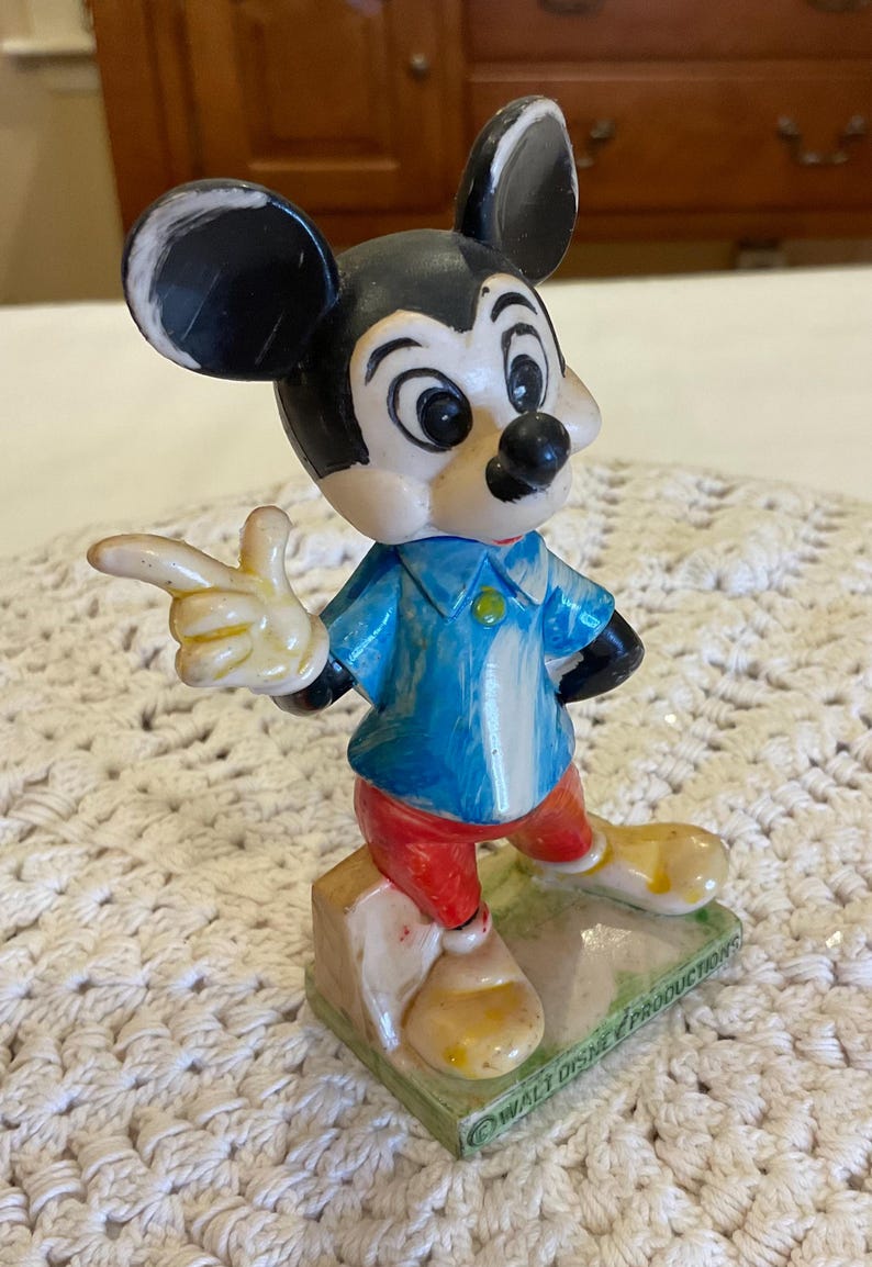 Peut inclure: Une figurine vintage en plastique de Mickey Mouse portant une chemise bleue, un pantalon rouge et des chaussures jaunes. La figurine est debout sur une base verte avec le texte "WALT DISNEY PRODUCTIONS" en bas.