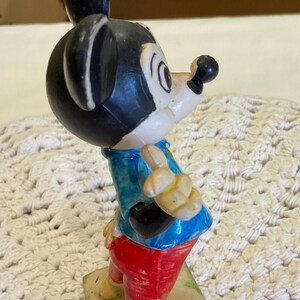 Peut inclure: Une figurine en plastique vintage de Mickey Mouse portant une chemise bleu et blanche, un pantalon rouge et des chaussures jaunes. La figurine est debout sur une base verte.