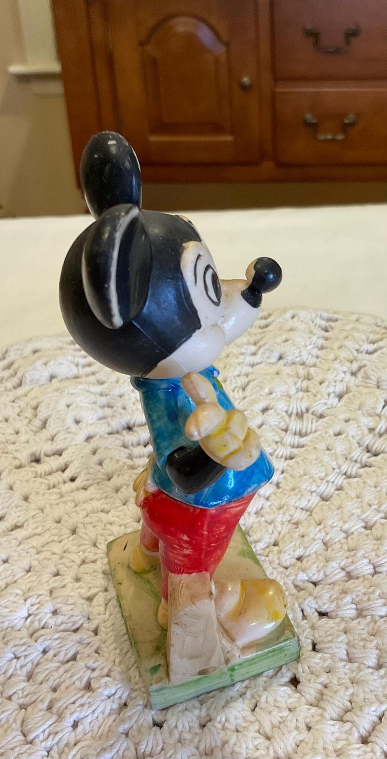 Peut inclure: Une figurine en plastique vintage de Mickey Mouse portant une chemise bleu et blanche, un pantalon rouge et des chaussures jaunes. La figurine est debout sur une base verte.