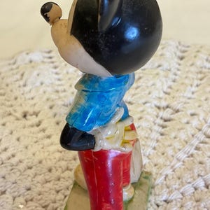 Peut inclure: Une figurine vintage de Mickey Mouse en plastique. Mickey porte une chemise &agrave; carreaux bleu et blanc, un pantalon rouge et des chaussures jaunes. Il est debout sur une base verte.