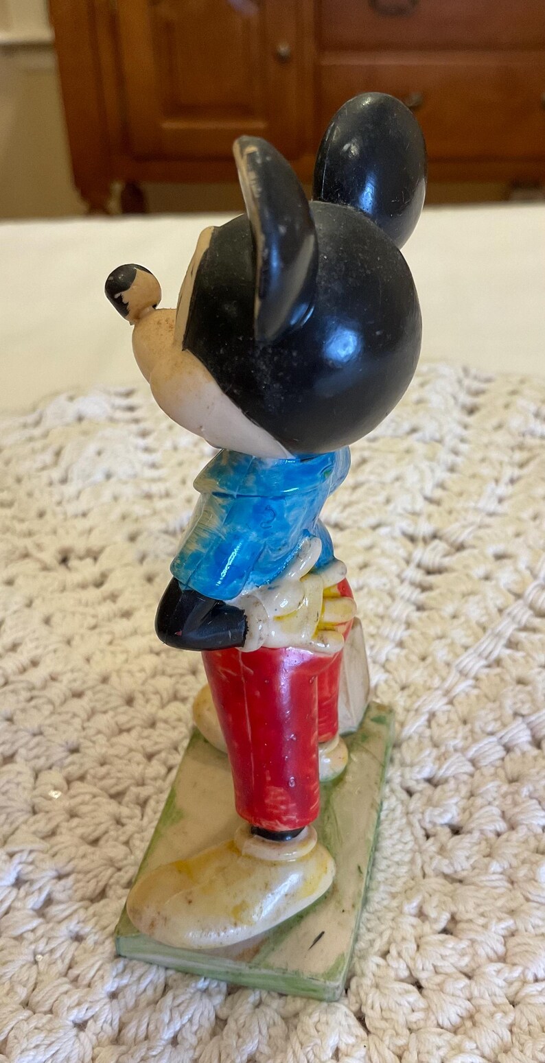 Peut inclure: Une figurine vintage de Mickey Mouse en plastique. Mickey porte une chemise &agrave; carreaux bleu et blanc, un pantalon rouge et des chaussures jaunes. Il est debout sur une base verte.