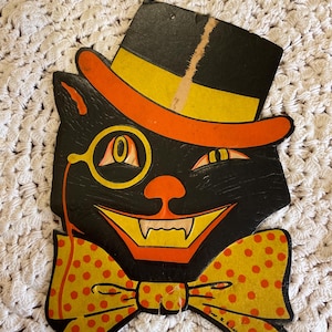 May include: Vintage Halloween decoration of a black cat wearing a top hat, bow tie, and monocle. The cat has a mischievous grin and yellow eyes. The hat is red and yellow, and the bow tie is yellow with orange polka dots. The text "Crowell & Littlejohn" is printed on the bottom of the decoration.