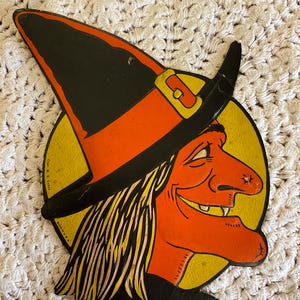 May include: Vintage Halloween decoration featuring a smiling witch's face. The witch has orange skin, a long nose, and yellow hair. She wears a black and orange witch hat with a gold buckle, set against a yellow circle.