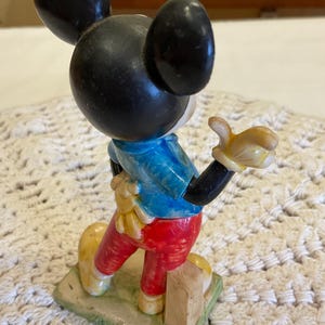 Peut inclure: Une figurine en plastique vintage de Mickey Mouse portant une chemise bleue, un pantalon rouge et des chaussures jaunes. Il est debout sur une base verte avec un bloc de bois brun.