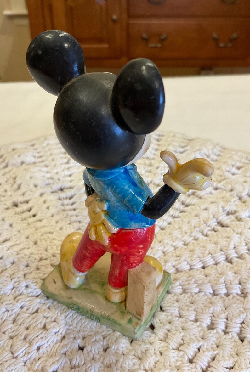 Peut inclure: Une figurine en plastique vintage de Mickey Mouse portant une chemise bleue, un pantalon rouge et des chaussures jaunes. Il est debout sur une base verte avec un bloc de bois brun.