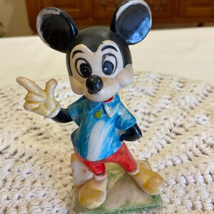 Peut inclure: Figurine en plastique vintage de Mickey Mouse pointant du doigt droit. Il porte une chemise ray&eacute;e bleu et blanche, un pantalon rouge et des chaussures jaunes. La figurine est debout sur une base verte avec le texte "Walt Disney Productions" dessus.