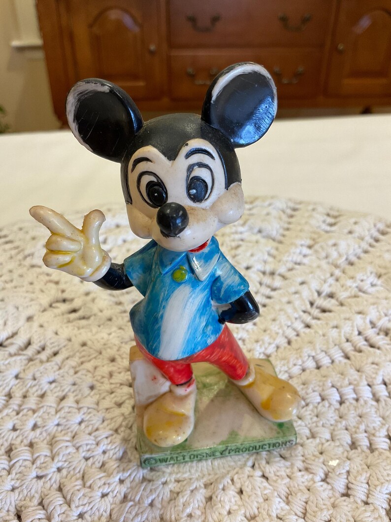 Peut inclure: Figurine en plastique vintage de Mickey Mouse pointant du doigt droit. Il porte une chemise ray&eacute;e bleu et blanche, un pantalon rouge et des chaussures jaunes. La figurine est debout sur une base verte avec le texte "Walt Disney Productions" dessus.