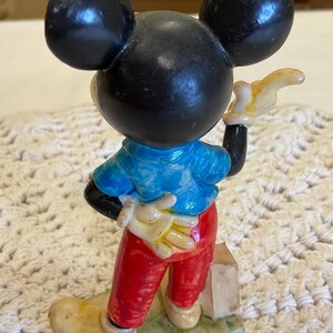 Peut inclure: Une figurine en c&eacute;ramique vintage de Mickey Mouse portant une chemise bleue, un pantalon rouge et des chaussures jaunes. Il tient un objet jaune dans sa main droite et a un visage noir et blanc avec de grandes oreilles.