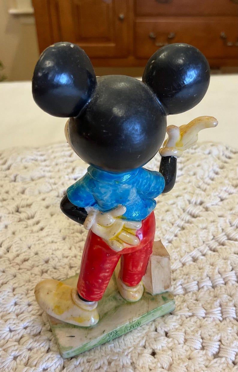 Peut inclure: Une figurine en c&eacute;ramique vintage de Mickey Mouse portant une chemise bleue, un pantalon rouge et des chaussures jaunes. Il tient un objet jaune dans sa main droite et a un visage noir et blanc avec de grandes oreilles.