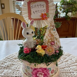 May include: A white ceramic bunny figurine with pink accents sits in a decorative planter with a white, pink, and green floral pattern. The planter is filled with green moss and artificial flowers. A small yellow duck figurine sits next to the bunny. A white sign with pink glitter and the text "Happy Spring" is attached to the planter.
