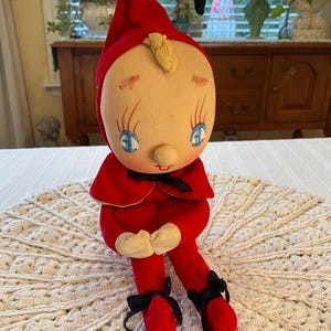 May include: A plush doll featuring a red pointed hat, a red cape with a black ribbon tie, and red pants. The doll has a beige face with blue eyes, long eyelashes, a small nose, and a single blonde hair curl. Black ribbons are tied around the ankles.