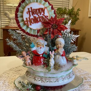 May include: A white ceramic bowl with a green rim and a green and brown floral pattern. The bowl is filled with faux snow and a Christmas scene with Santa Claus, a woman in a red dress, a white rabbit, a blue bird, and a cherub. The bowl is sitting on a white crocheted doily. A red and green paper fan with the words "Happy Holidays" is in the center of the arrangement.