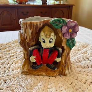 May include: A ceramic planter shaped like a tree trunk with a small gnome figure sitting inside. The gnome is wearing a red and black outfit and has a white face with brown hair. The planter is decorated with a purple flower and green leaves.