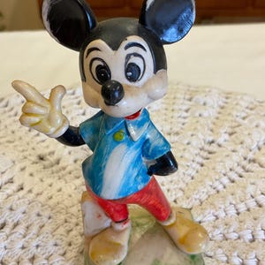 Peut inclure: Figurine vintage de Mickey Mouse debout sur une base verte avec le texte "&copy; WALT DISNEY PRODUCTIONS". Mickey porte une chemise bleu et blanche, un pantalon rouge et des chaussures jaunes. Il pointe du doigt avec sa main droite.