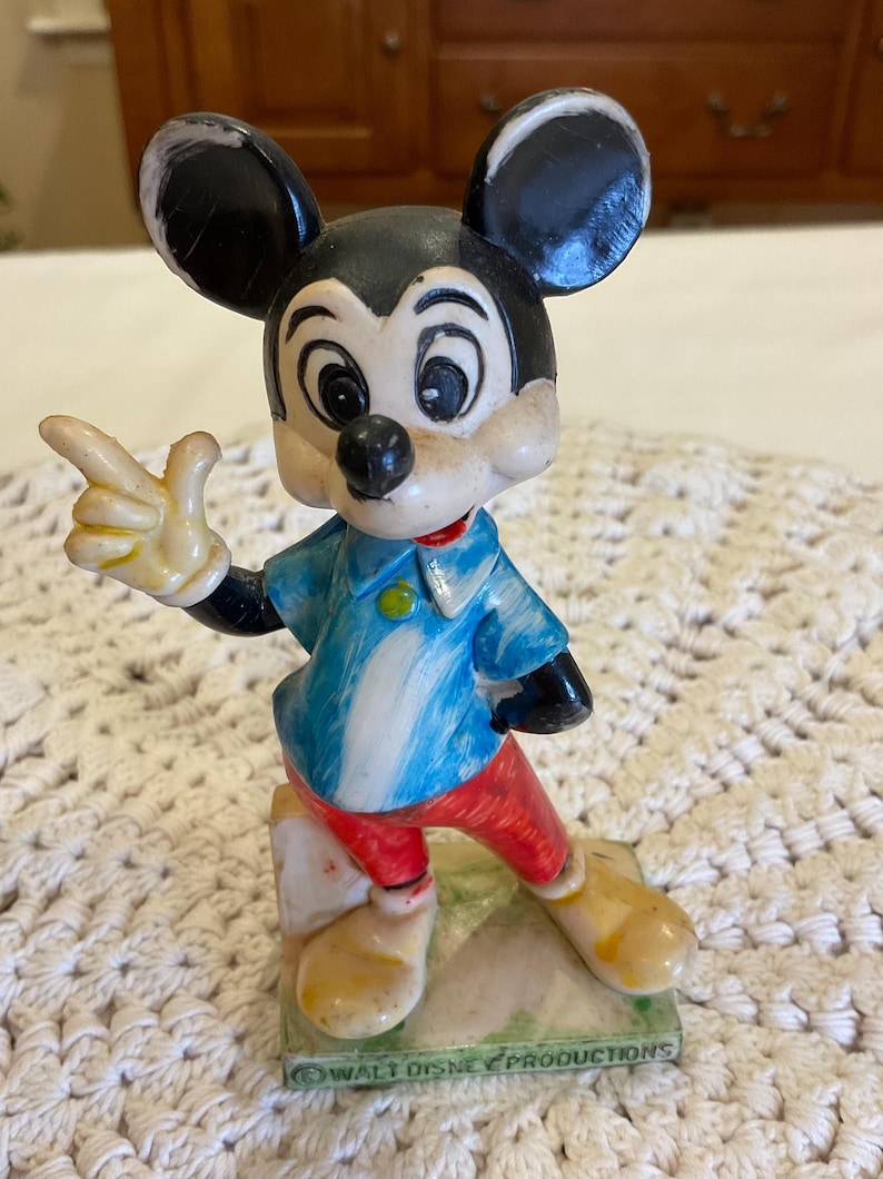 Peut inclure: Figurine vintage de Mickey Mouse debout sur une base verte avec le texte "&copy; WALT DISNEY PRODUCTIONS". Mickey porte une chemise bleu et blanche, un pantalon rouge et des chaussures jaunes. Il pointe du doigt avec sa main droite.