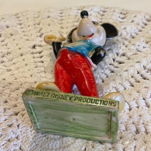 Peut inclure: Figurine vintage de Mickey Mouse avec un pantalon rouge, une chemise bleue et des oreilles noires. La figurine est debout sur une base verte et blanche avec le texte "&copy; WALT DISNEY PRODUCTIONS" au bas.