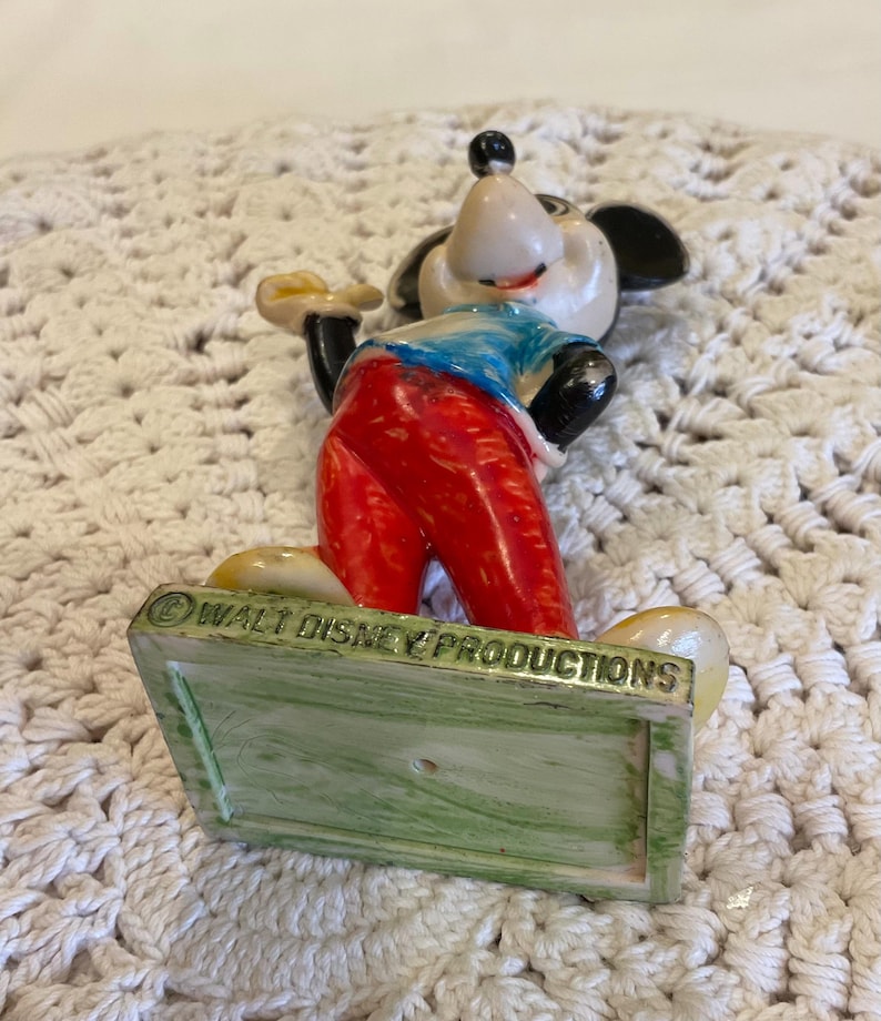 Peut inclure: Figurine vintage de Mickey Mouse avec un pantalon rouge, une chemise bleue et des oreilles noires. La figurine est debout sur une base verte et blanche avec le texte "&copy; WALT DISNEY PRODUCTIONS" au bas.