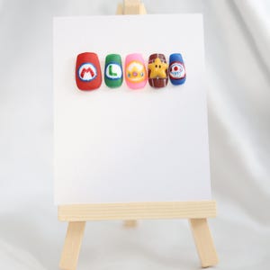 May include: Five colourful nail art designs displayed on a white card, resting on a small wooden easel. The designs feature cartoon characters and letters, including a red nail with a white "M", a green nail with a white "L", and a pink nail with a yellow character.