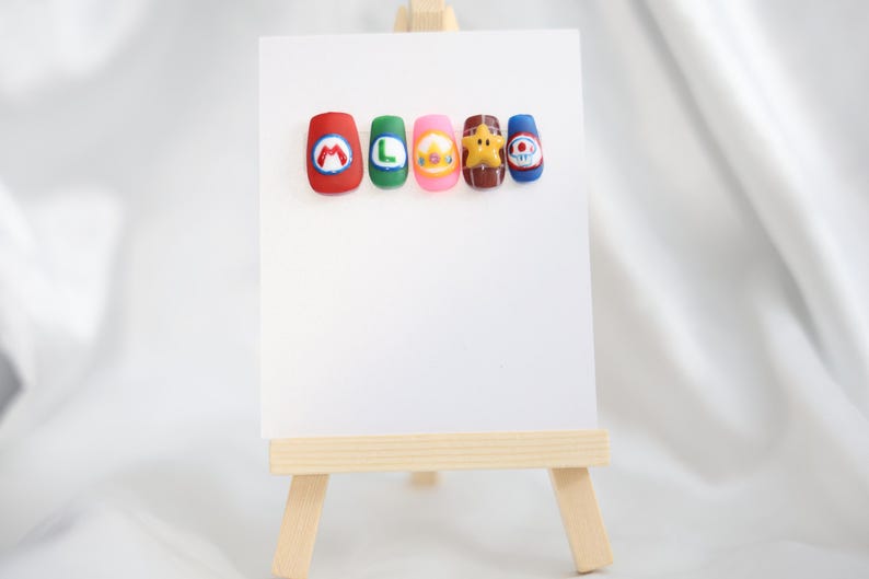 May include: Five colourful nail art designs displayed on a white card, resting on a small wooden easel. The designs feature cartoon characters and letters, including a red nail with a white "M", a green nail with a white "L", and a pink nail with a yellow character.