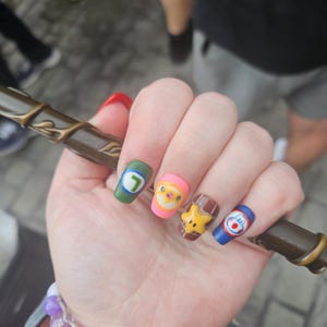 May include: A hand with decorated nails, each with a different cartoon character design. The nails are painted in various colours, including green, pink, brown, and blue. The hand is holding a brown wand. A purple and white beaded bracelet is on the wrist.