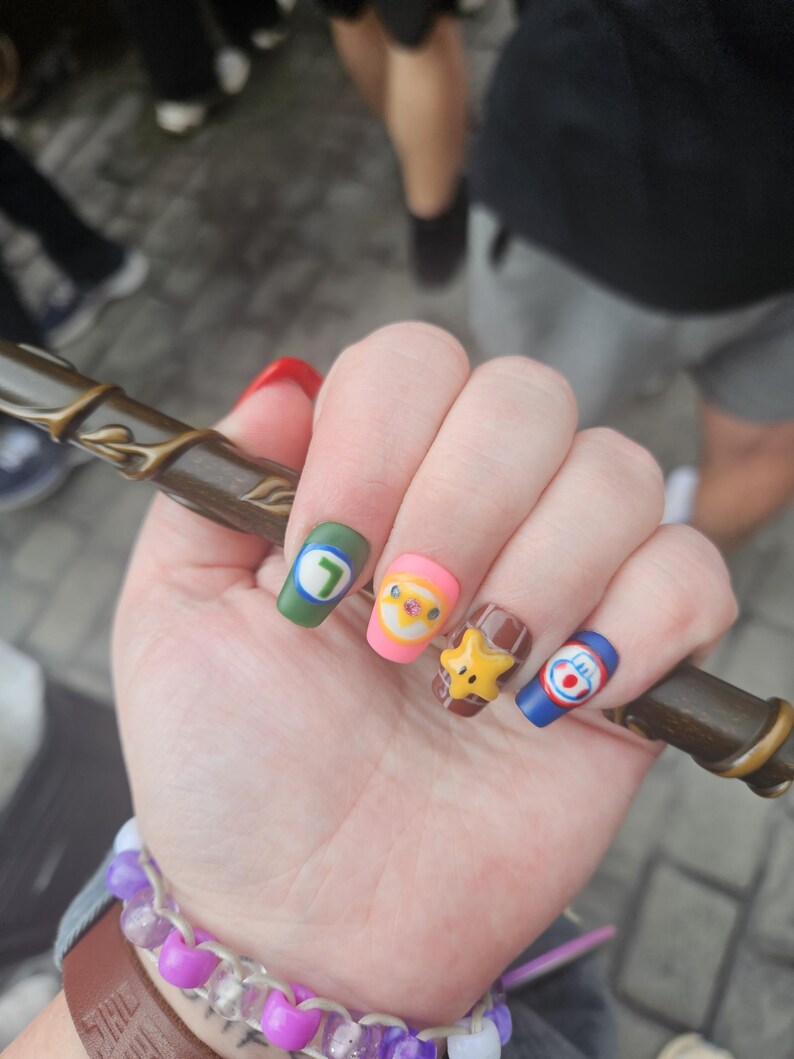 May include: A hand with decorated nails, each with a different cartoon character design. The nails are painted in various colours, including green, pink, brown, and blue. The hand is holding a brown wand. A purple and white beaded bracelet is on the wrist.