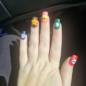 May include: A hand with long, colourful, cartoon-themed nail art. Each nail features a different design, including a star, a character with a crown, and the letters "L" and "M". The nails are painted in various colours, including red, blue, green, and pink.