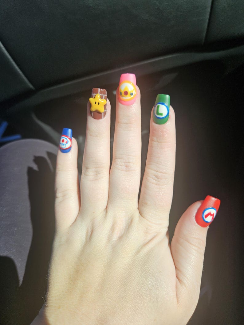 May include: A hand with long, colourful, cartoon-themed nail art. Each nail features a different design, including a star, a character with a crown, and the letters "L" and "M". The nails are painted in various colours, including red, blue, green, and pink.