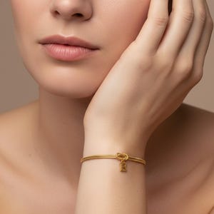 Op de afbeelding: Goudkleurige armband met een knoopontwerp en een kleine letter "E" bedel. De armband, gedragen om de pols, is gemaakt van dun, getextureerd metaal. De achtergrond is een neutraal beige.