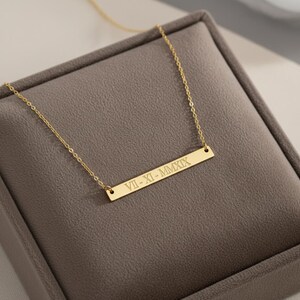 May include: A gold-colored bar necklace with a delicate chain. The rectangular pendant is engraved with the Roman numerals VII-XI-MMXIX. The necklace is displayed on a gray velvet surface.